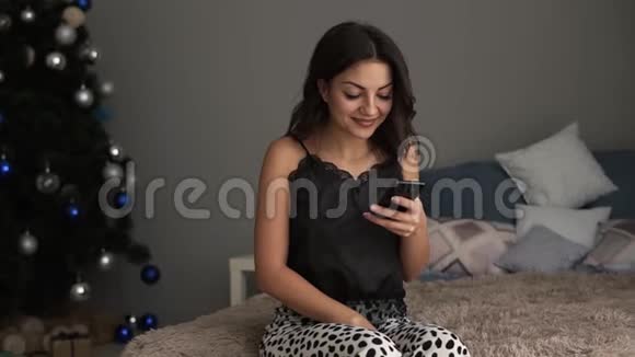 快乐的黑发女人坐在床上看着她的手机美丽的女孩收到圣诞礼物视频的预览图