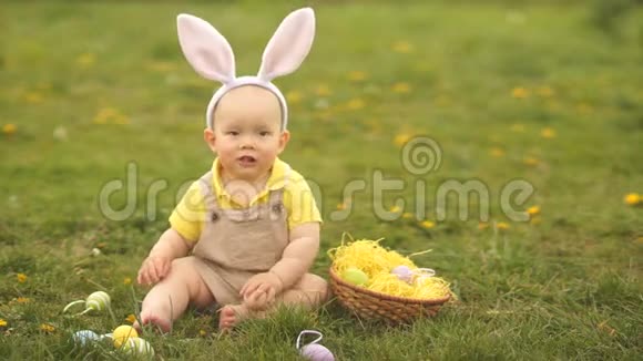 可爱的宝宝穿着复活节兔子的服装坐在公园的草地上的篮子里收集复活节彩蛋春季野餐视频的预览图