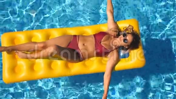 漂亮的女孩戴着太阳镜和红色比基尼躺在游泳池里的黄色充气床垫上年轻晒黑的女人视频的预览图