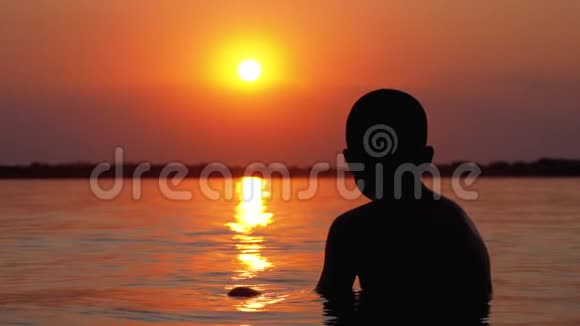 日落和橙径背景下坐在水里的男孩剪影慢动作视频的预览图