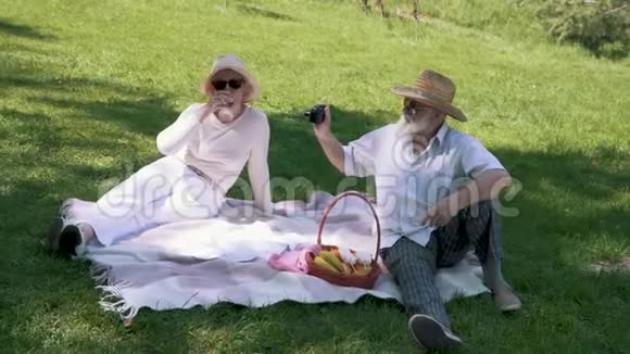 戴着帽子的成熟男人在野餐时用一杯酒为他优雅的妻子拍照视频的预览图