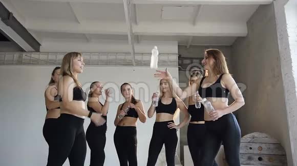 年轻的运动女性在训练后喝着瓶子的水同时站在健身房的白色墙壁上女性视频的预览图