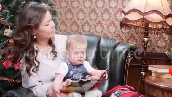 一位年轻的母亲和一个小儿子坐在圣诞树旁的皮椅上看着一本儿童书视频的预览图