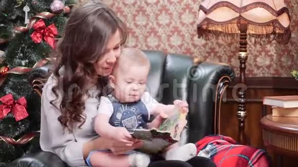 一位年轻的母亲和一个小儿子坐在圣诞树旁的皮椅上看着一本儿童书视频的预览图