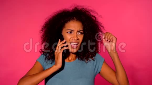 愤怒的非洲裔美国妇女一头卷发穿着绿色t恤在粉色墙背景下用智能手机打电话视频的预览图