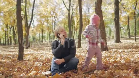 一个女人坐在秋天公园里打电话一个小女儿在附近玩视频的预览图