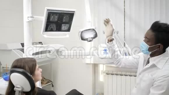 打开牙科灯年轻的非裔美国男医生戴着医疗面具的牙医坐在椅子上的漂亮女病人视频的预览图