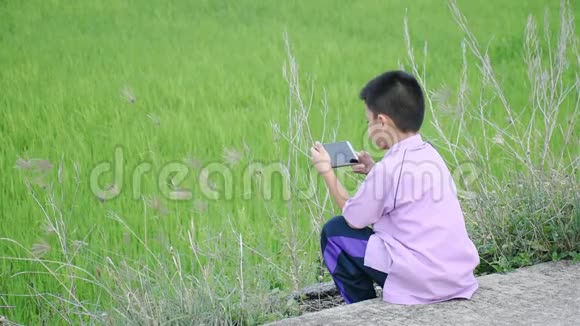 一个亚洲男孩拿着电话坐在街上绿色的稻田视频的预览图