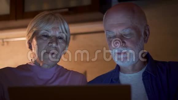 家里的老夫妇晚上在厨房里用笔记本电脑退休养恤金领取者的家庭在电脑上工作视频的预览图