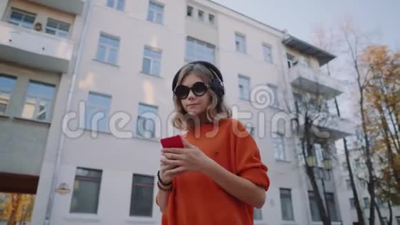 可爱的少女听耳机音乐都市风格时髦的时髦少女选择手机轨道微笑视频的预览图