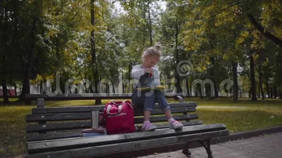 小美丽的学校女孩用彩色铅笔画画坐在阳光明媚的公园里的长凳上慢动作镜头视频的预览图
