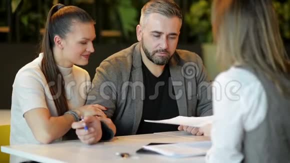 坐在办公室桌上讨论出售房产的房地产经纪人和年轻夫妇的侧视图幸福的夫妻成了房子视频的预览图