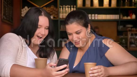 两个兴奋超重的白种人女孩在咖啡馆里打电话喝咖啡视频的预览图
