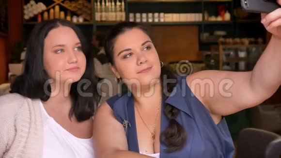 两个肥胖的白种人女孩坐在咖啡馆里用智能手机拍照视频的预览图