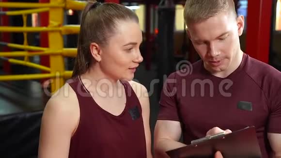 肌肉型个人教练在剪贴板上写作并在交叉训练健身房向年轻女性讨论训练计划视频的预览图