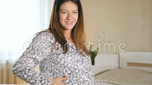 怀孕的年轻女人看着她圆圆的肚子抚摸着它卧室床上穿着滑稽睡衣的女孩视频的预览图