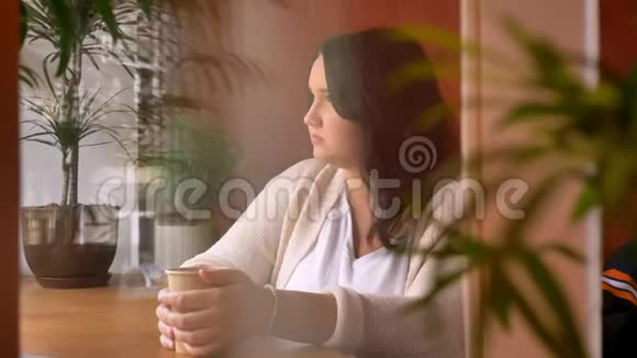 一个体贴入微的白种人女孩从纸杯里喝酒看着窗户的室内视频的预览图