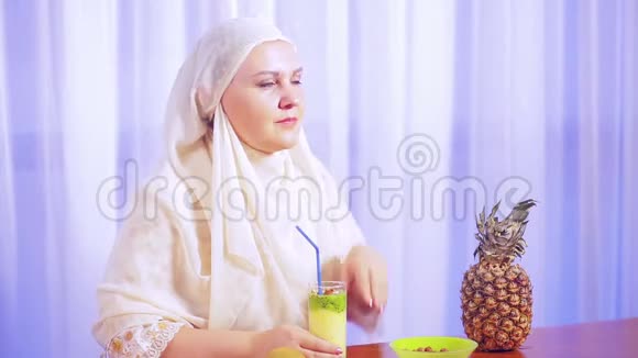 一位年轻的穆斯林妇女戴着浅色围巾喝着水果奶昔微笑着视频的预览图