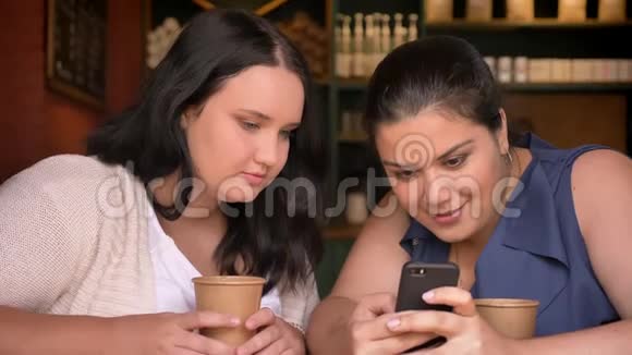 两个白种人的大女人一起在智能手机上刷卡室内还有一杯咖啡在桌子上视频的预览图