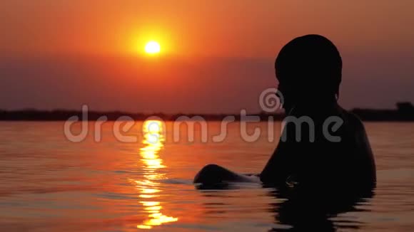 日落和橙色小径背景下坐在水里的男孩剪影慢动作视频的预览图