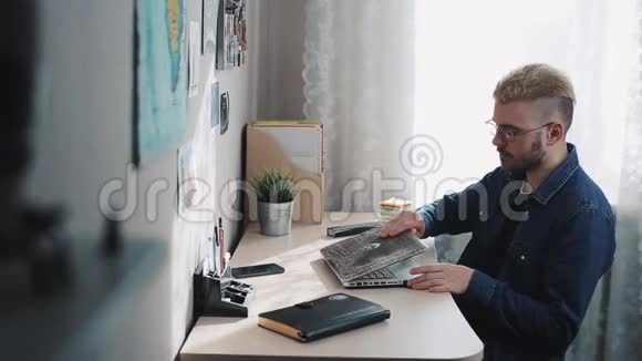 年轻的男性家庭自由职业者眼镜和黄色头发打开和开始使用笔记本电脑家庭工作场所内政部视频的预览图