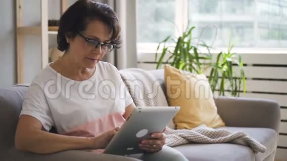 穿白色T恤的成熟女人坐在家里的沙发上视频的预览图