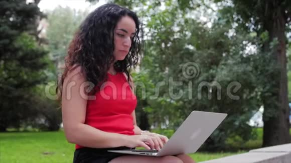 在笔记本电脑上工作的女人穿着黑色短裤的漂亮的布鲁内特在公园里做着笔记本电脑的工作女孩在她的身上打印出来视频的预览图