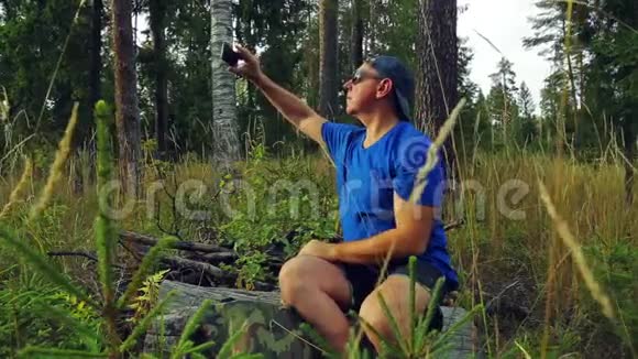 一位戴着太阳镜和棒球帽的男游客坐在一棵倒下的树上在电话里拍照视频的预览图