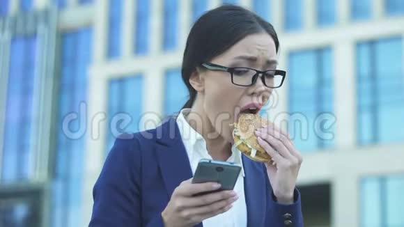 年轻女经理匆匆吃着不健康的汉堡在电话里查看新闻视频的预览图