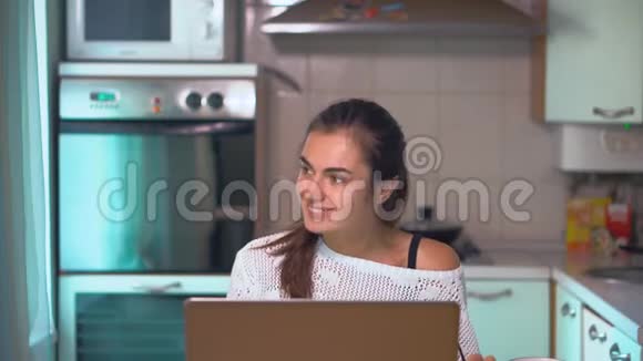白人黑发女孩坐在厨房里上网学习并在笔记本电脑上搜索信息视频的预览图