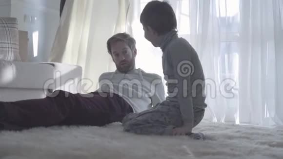 小胡子爸爸和孩子说话大胡子爸爸躺在白色地毯上儿子坐在他旁边父亲视频的预览图