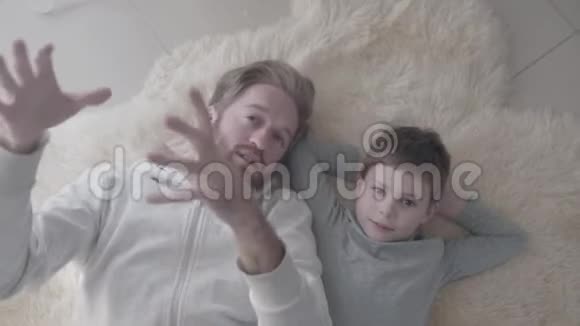 大胡子爸爸和他的小儿子躺在白色的毛皮地毯上进行有趣的情感对话父亲子女视频的预览图