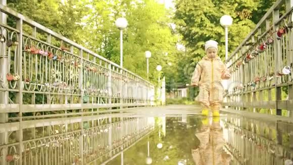穿着橙色防水雨衣和橡胶雨靴的滑稽小女孩在雨后的水坑里慢动作视频的预览图