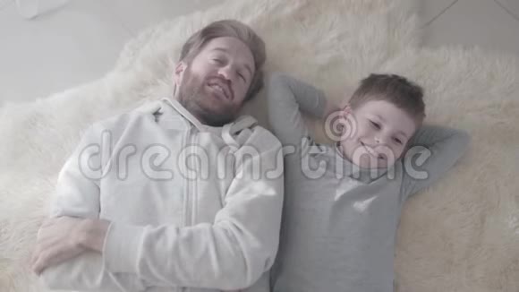 快乐的微笑爸爸和他快乐的儿子躺在毛皮地毯上父子关系父子关系视频的预览图