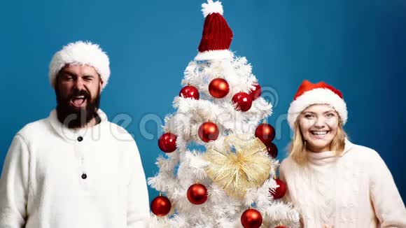 年轻夫妇在蓝底圣诞树附近做鬼脸有胡子的男人和金发女人站在树旁视频的预览图