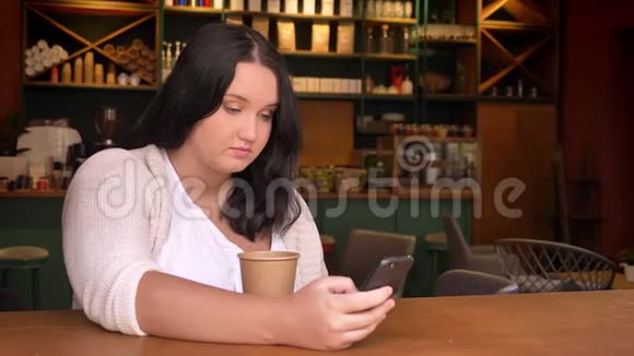 在咖啡屋里端着一杯饮料坐着的时候白种人中的一些腐烂的女人在拿着智能手机视频的预览图