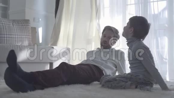 父亲和孩子说话大胡子爸爸躺在白色地毯上儿子坐在他旁边父亲子女视频的预览图