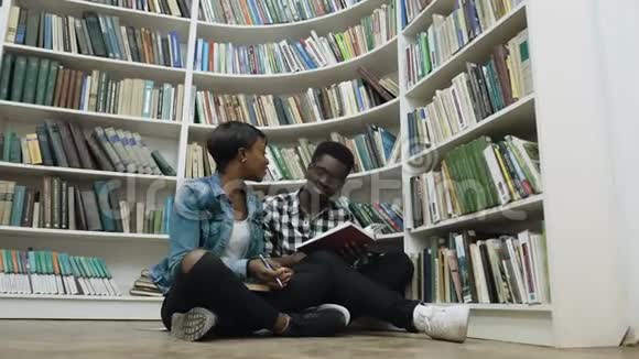 年轻的非洲夫妇在图书馆准备考试时坐在书架附近的地板上视频的预览图