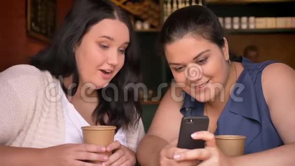 两个多肉的白种人女孩在咖啡馆里拿着纸杯在感情上讨论着看电话视频的预览图