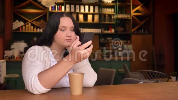 沉重的白种人女性手里拿着手机坐在咖啡馆里手里拿着一个大纸杯视频的预览图