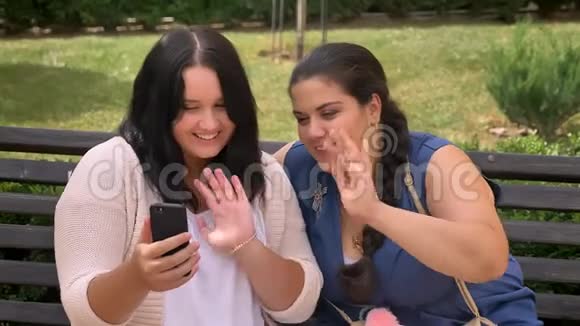 两个年轻的肥胖女孩在智能手机上对着镜头挥手在公园的长椅上放松地微笑视频的预览图