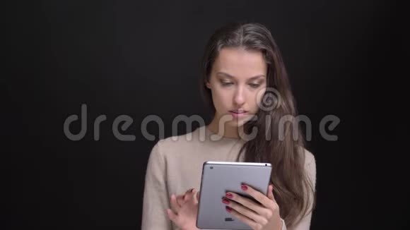 年轻的黑发白种人长发女孩的肖像微笑地看着黑背景的平板电脑视频的预览图