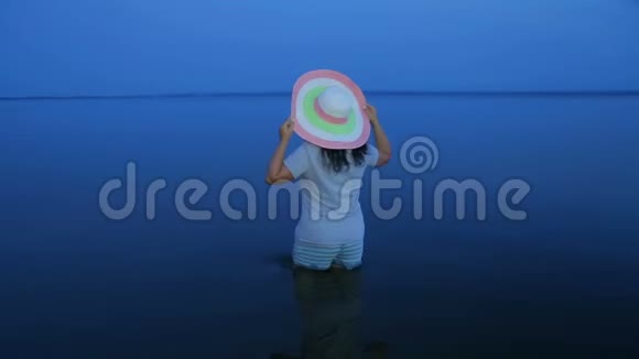 一个穿着短裤和帽子的年轻微笑的女人站在湖边的膝盖上欣赏日落视频的预览图