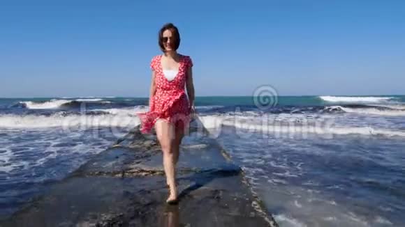 年轻的微笑快乐的女人赤脚走在海上码头上红色连衣裙随风飘扬波浪撞击海港视频的预览图