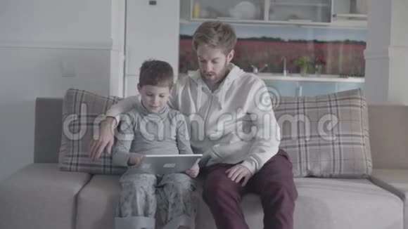 父亲和孩子坐在大客厅的沙发上在平板电脑屏幕上滚动爸爸教他的孩子父子关系视频的预览图