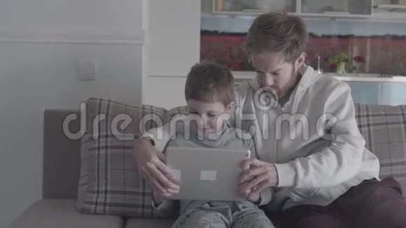 微笑快乐的父亲和孩子使用数字平板电脑坐在沙发上的大客厅爸爸教儿子视频的预览图