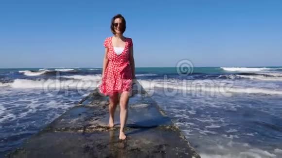 年轻的微笑快乐的女人赤脚走在海上码头上红色连衣裙随风飘扬波浪撞击海港慢动作视频的预览图