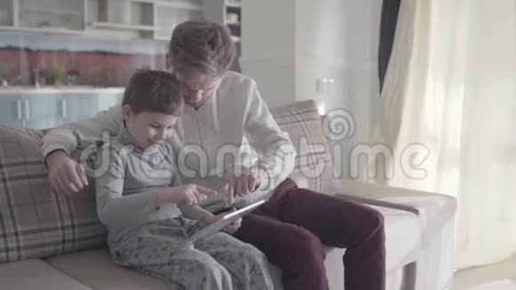 父亲和孩子坐在大客厅的沙发上在平板电脑屏幕上滚动爸爸教他的孩子父子关系视频的预览图