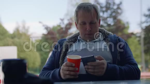 一个穿着t恤手里拿着纸杯咖啡的人在智能手机上上网视频的预览图