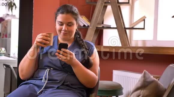超大尺寸的白种人女士喝着咖啡坐在沙发上滚动着她的智能手机视频的预览图